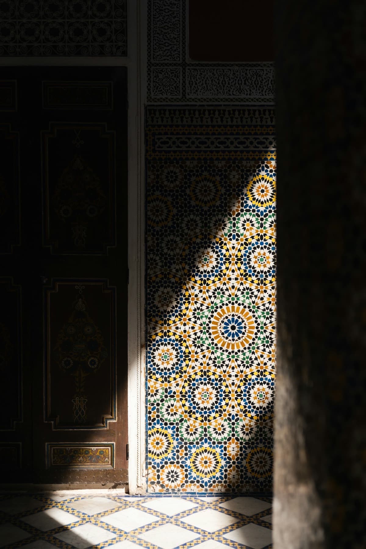 Light falling across a wall of hand-cut zellige in a Moroccan riad, beside a carved and painted door.